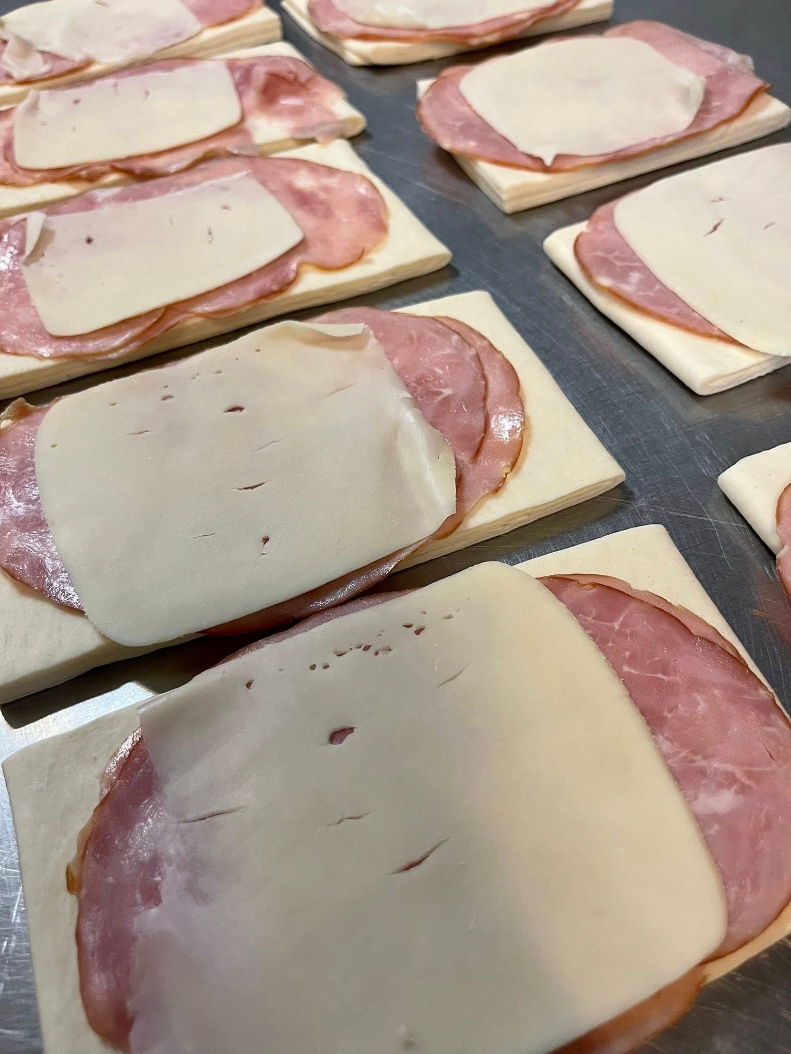 Sandwiches on rectangular bread with meat and cheese, prepared on a metal surface.