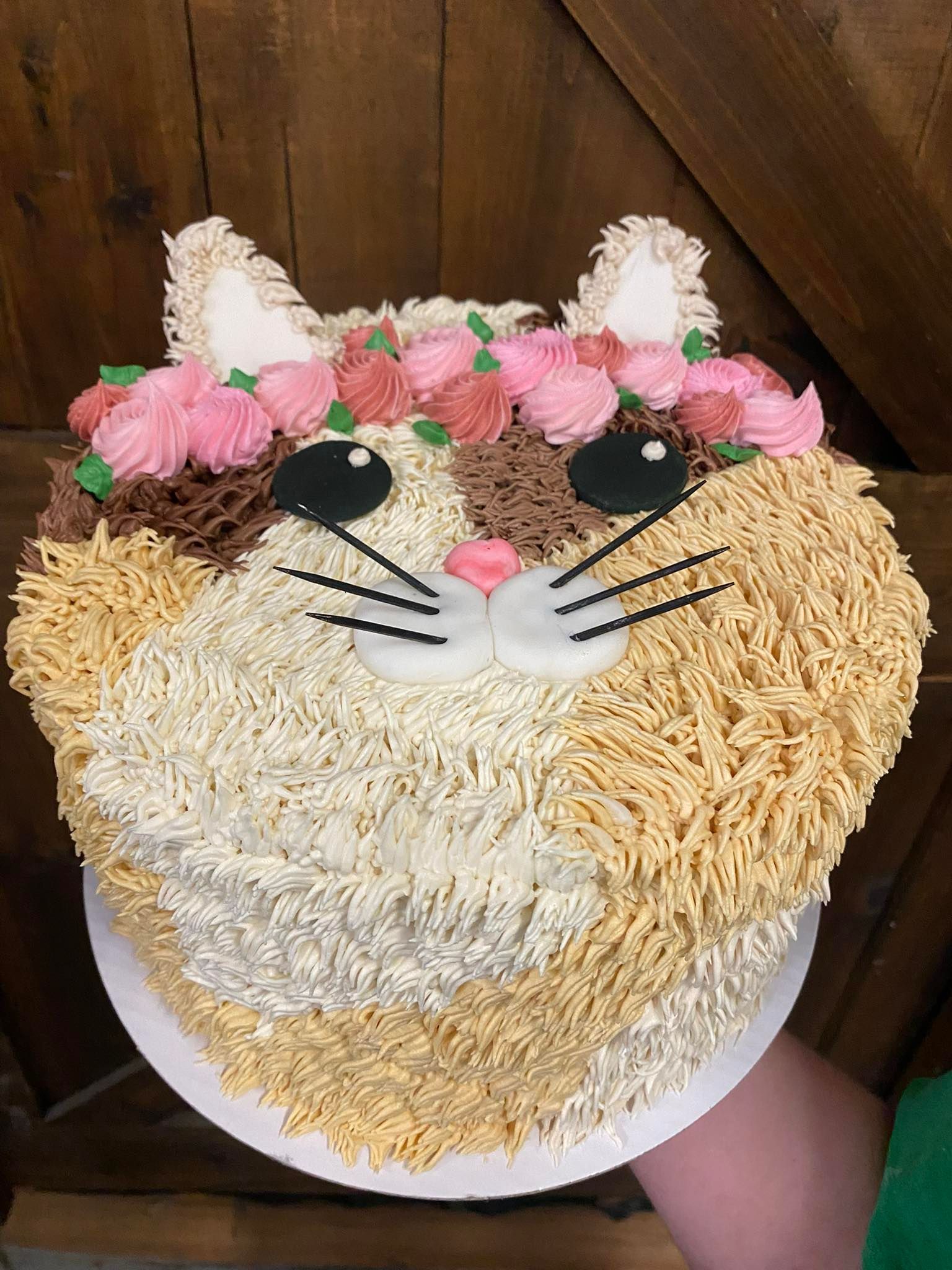 Cat-shaped cake, decorated with tan, white, brown frosting. It has a flower crown, black eyes, whiskers, and a pink nose.