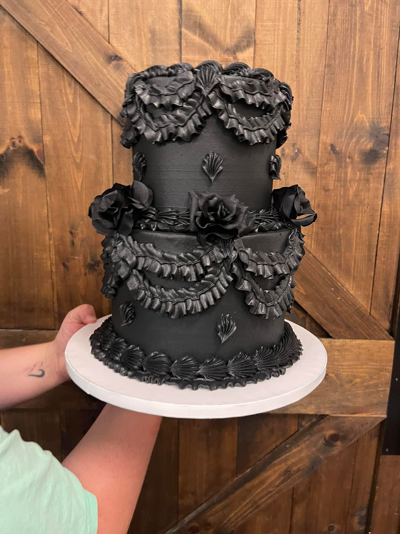Two-tiered black cake with ornate frosting detail, held by person on a white plate, against a wooden background.