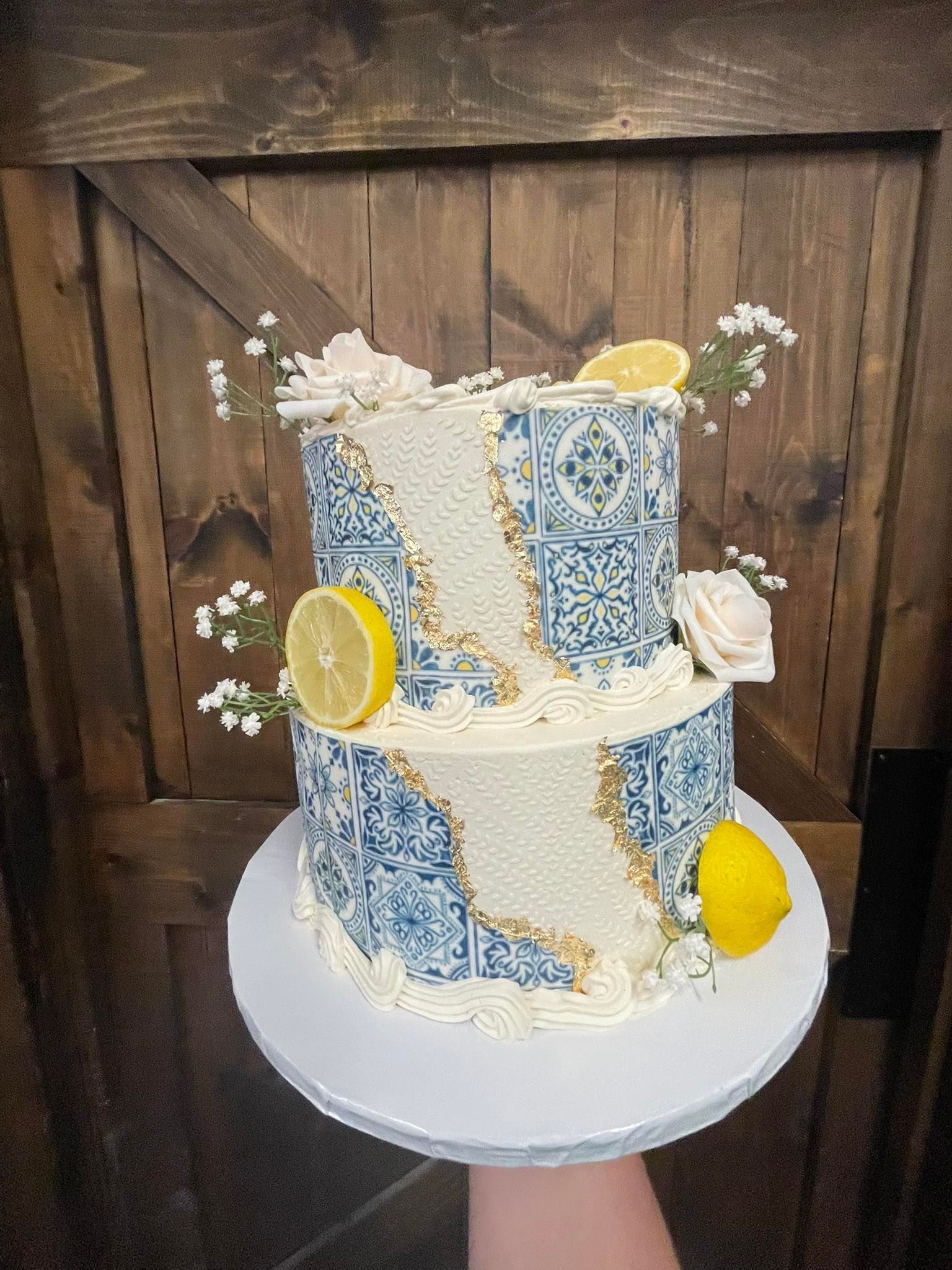 Two-tiered cake with blue and white tile design, decorated with lemons, flowers, and gold accents.
