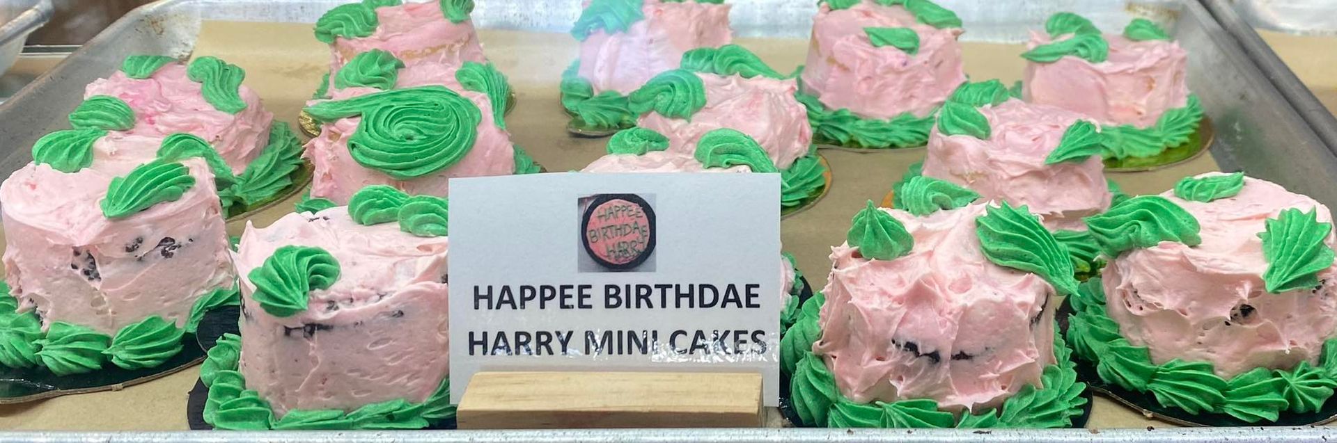 Mini cakes with pink frosting and green decorations arranged on a tray. A sign says 