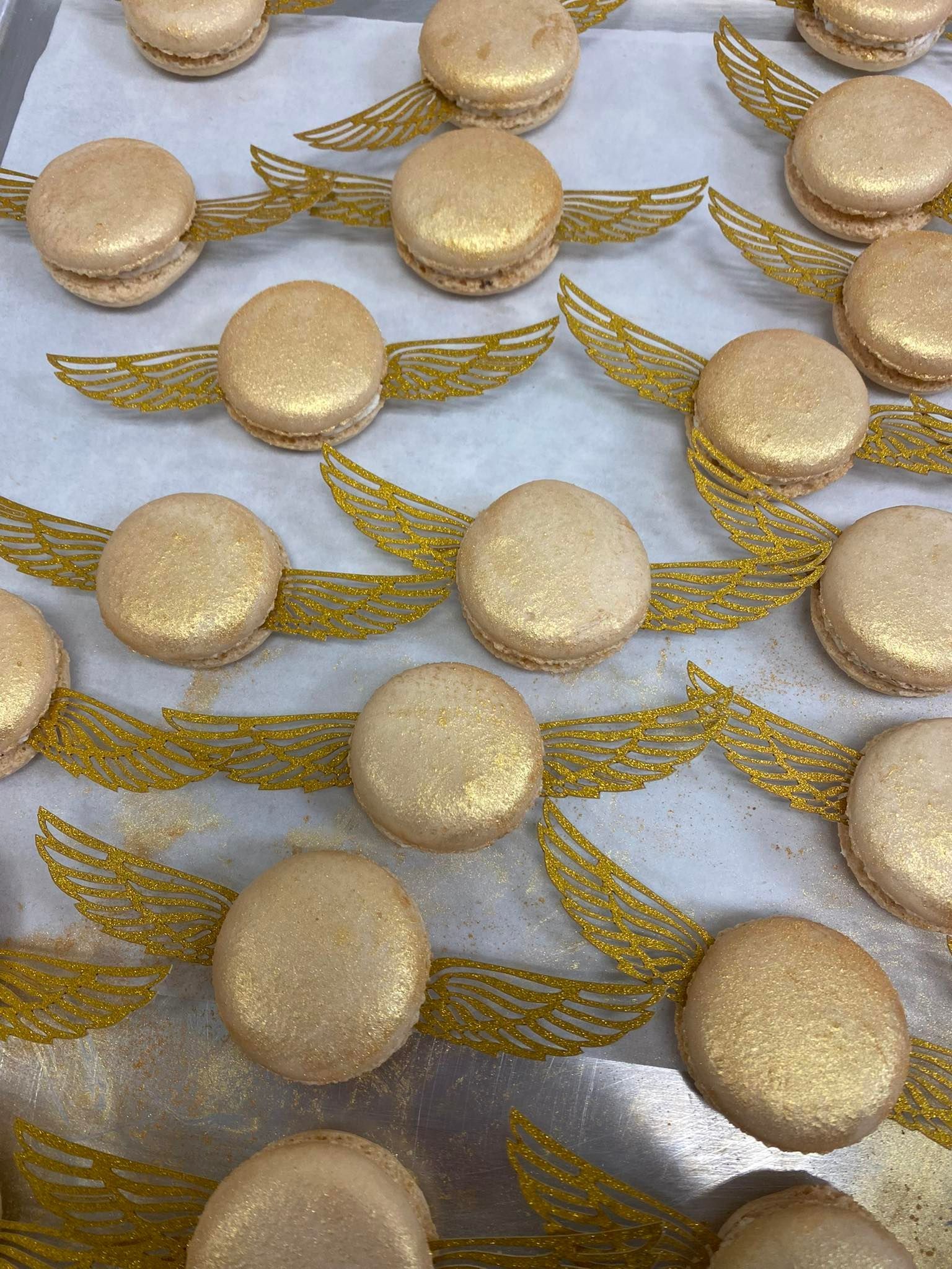 Golden macarons with glitter, decorated with gold wings, on a tray.