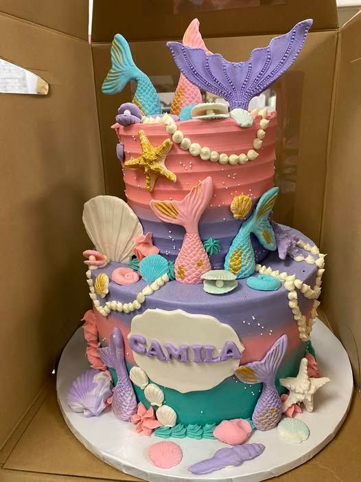 Two-tiered mermaid cake with teal, purple, and pink frosting, seashell and pearl decorations, and mermaid tail accents.