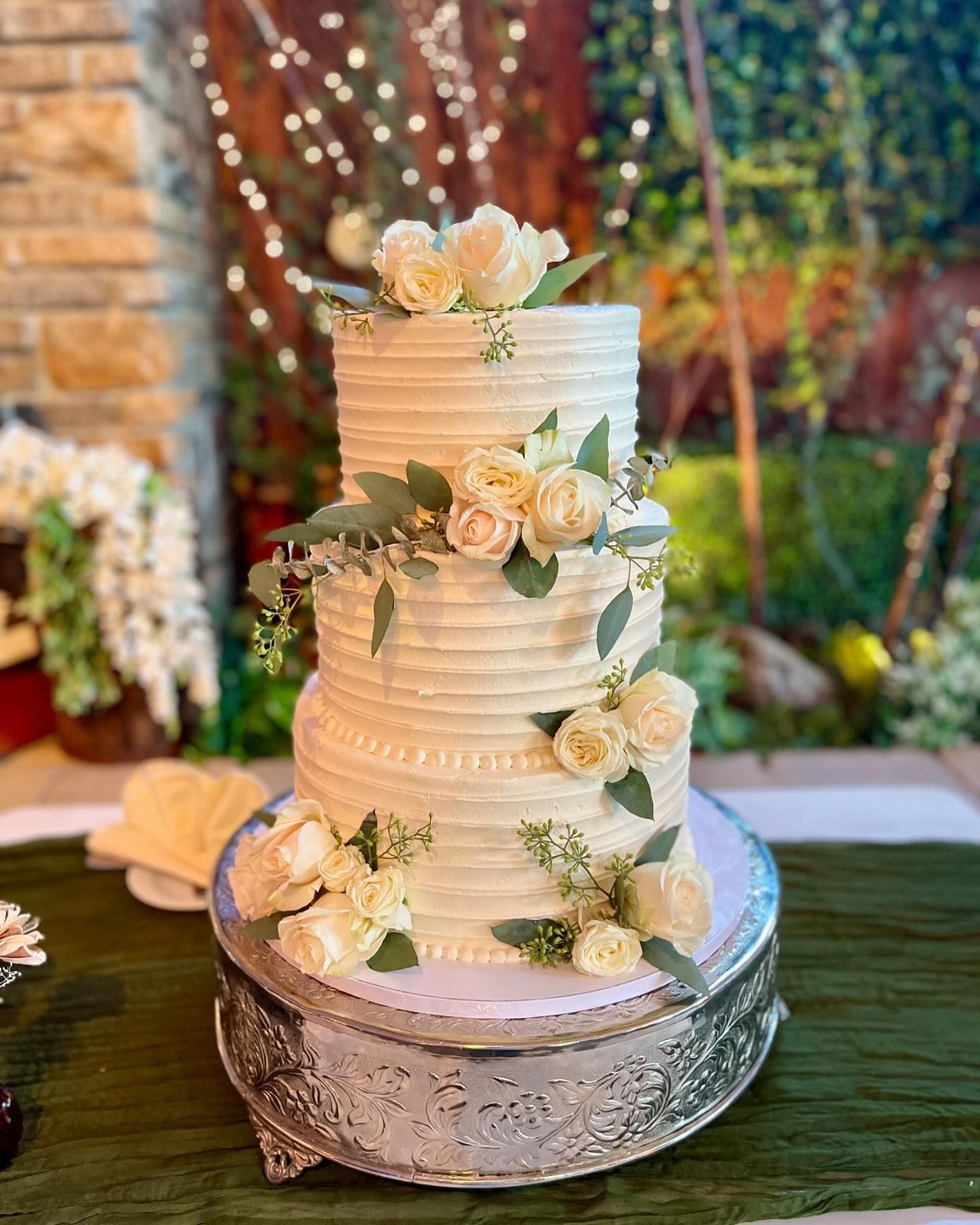 Three-tiered wedding cake with white frosting and floral decorations on a silver stand, set on a green table.