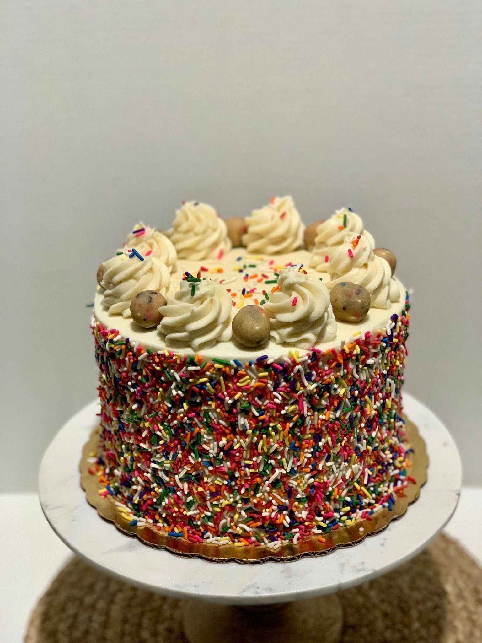 A round cake with rainbow sprinkles and swirled frosting, adorned with small cookie balls, on a white cake stand.