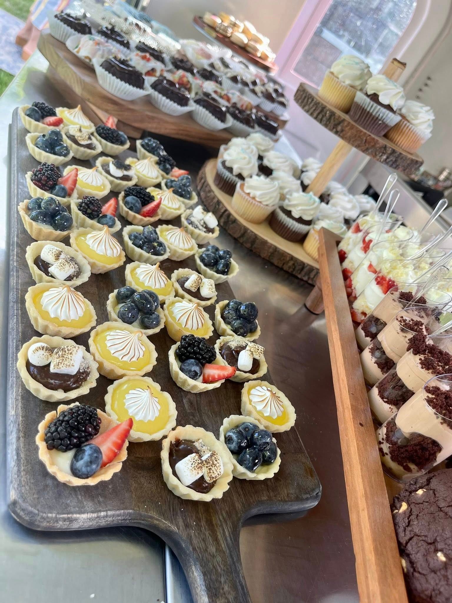 Dessert table with mini tarts, cupcakes, and chocolate brownies, outdoors on a sunny day.