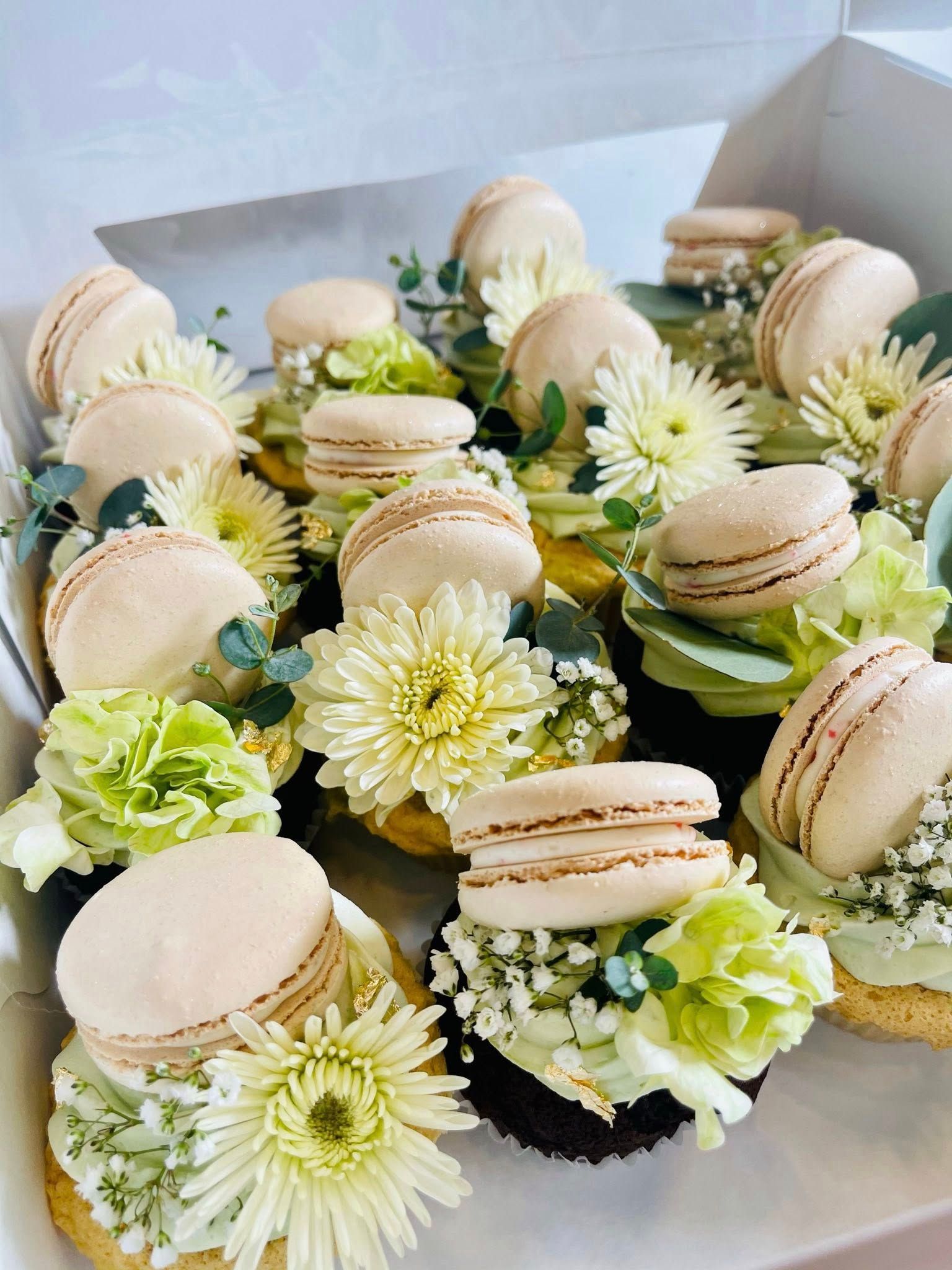 Box of cupcakes with light brown macarons, yellow and green flowers.