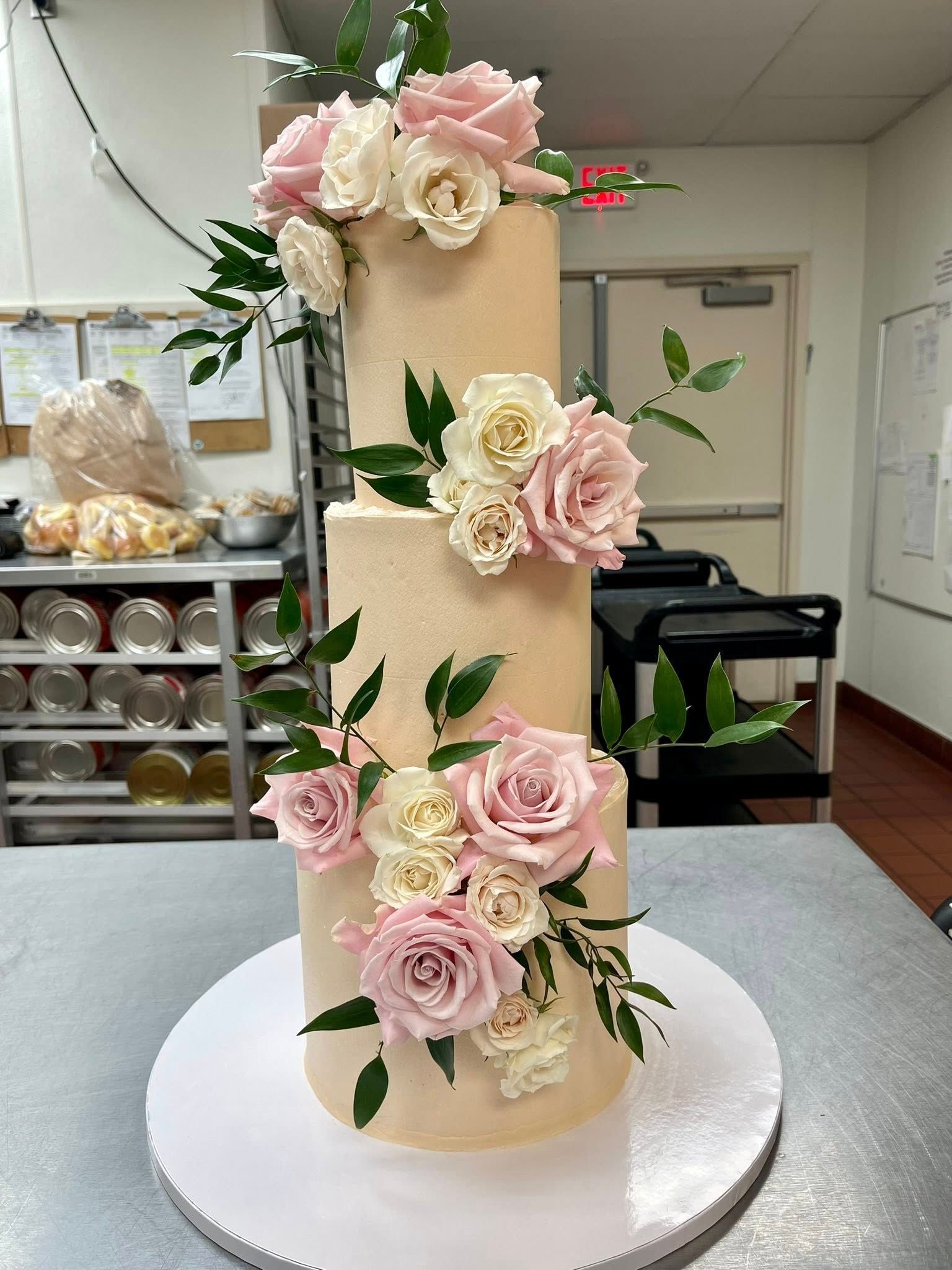 Three-tier cake with light pink frosting, decorated with pink and white roses and greenery, on a white cake board.