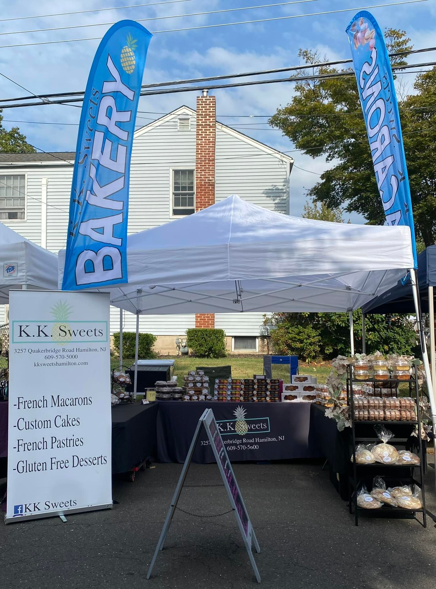 Bakery stand with blue flags and white tent, selling baked goods. Display sign in front of a white house.