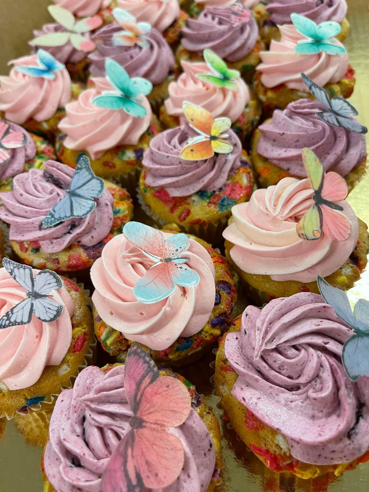 Cupcakes with pastel-colored frosting and butterfly decorations, arranged on a gold surface.