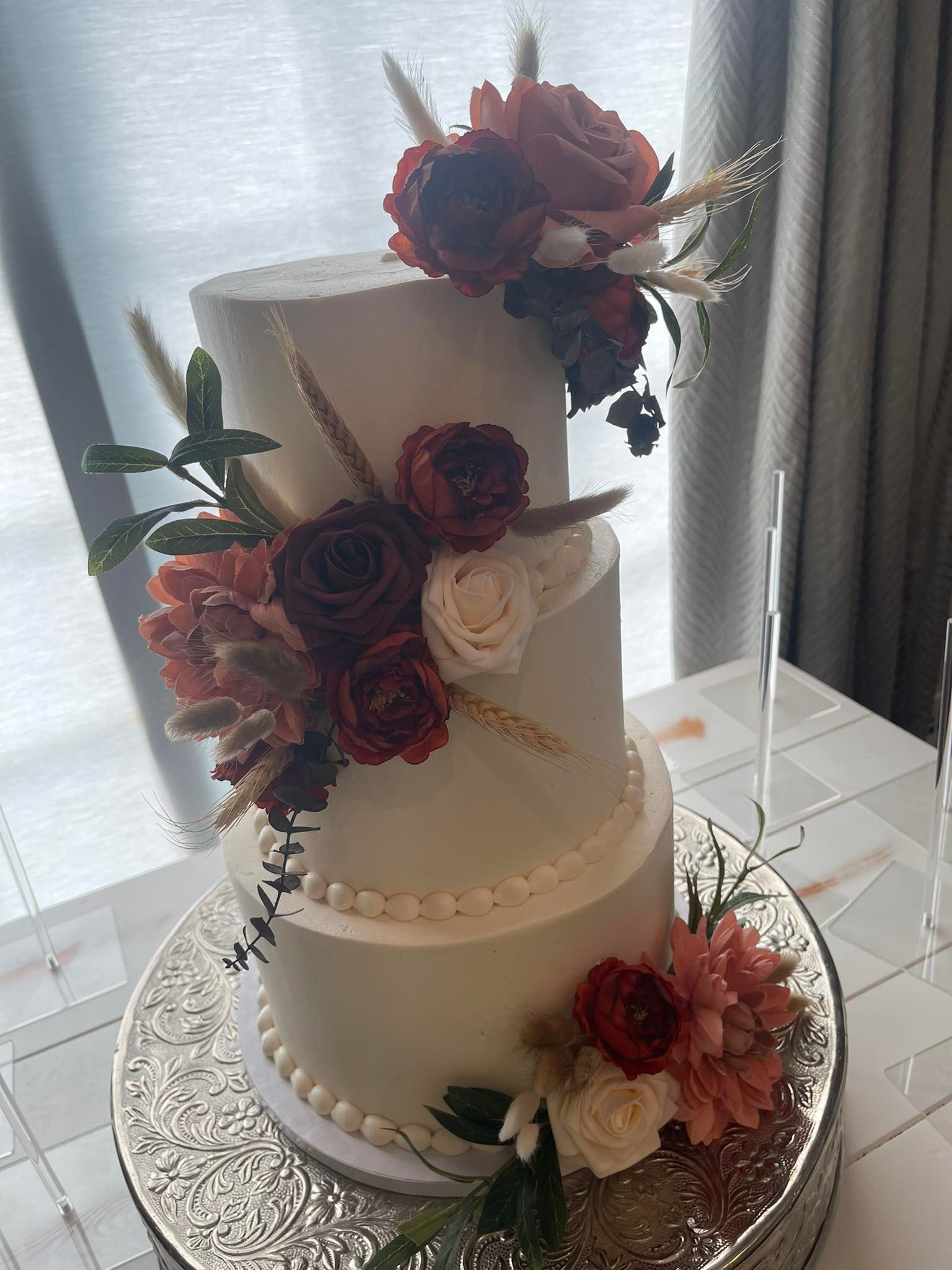 Three-tiered cake with white frosting and burgundy, peach, and white flowers, on a silver cake stand.