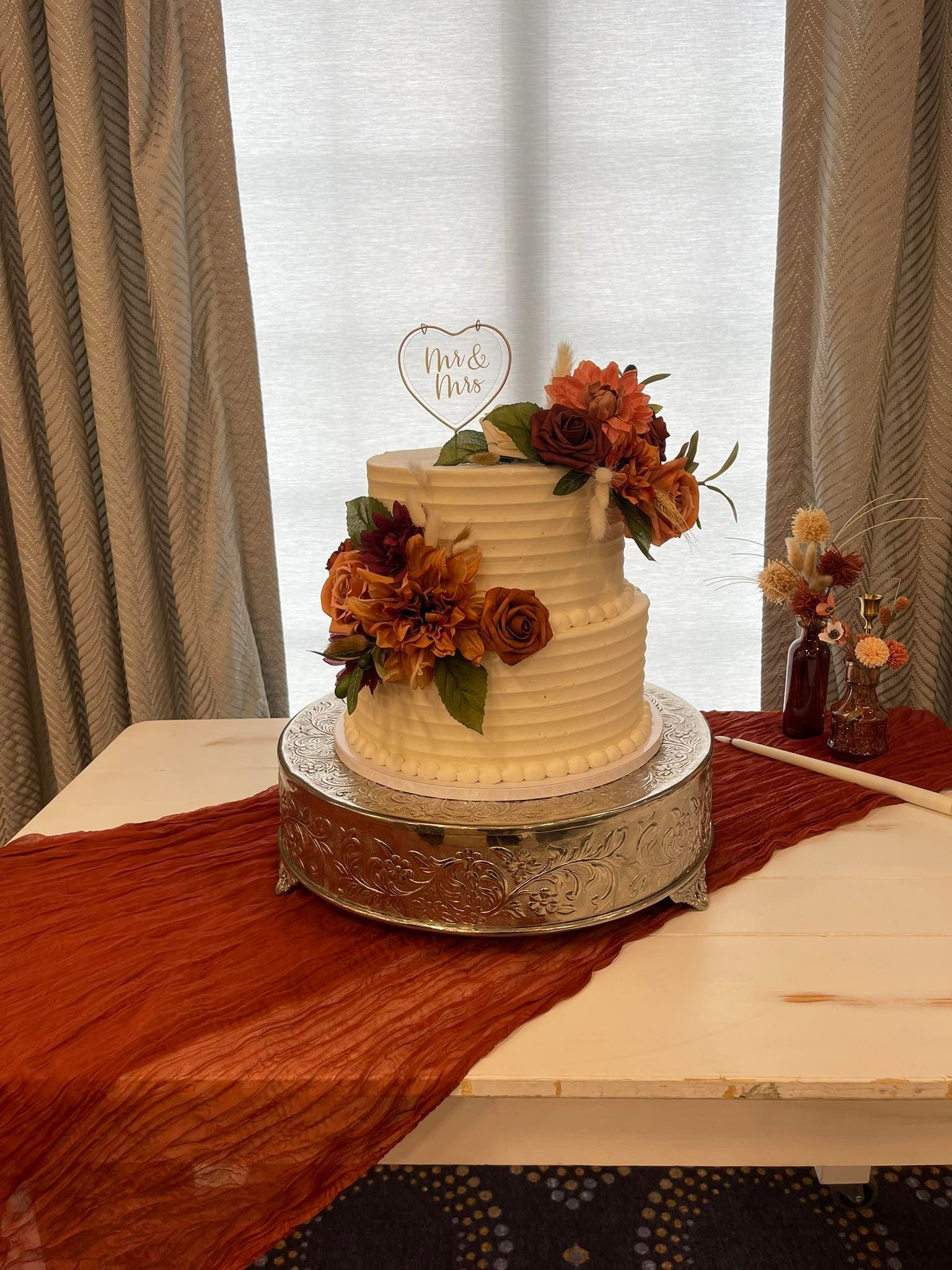 Two-tiered wedding cake with orange flowers, heart topper, on a silver stand, and red runner.