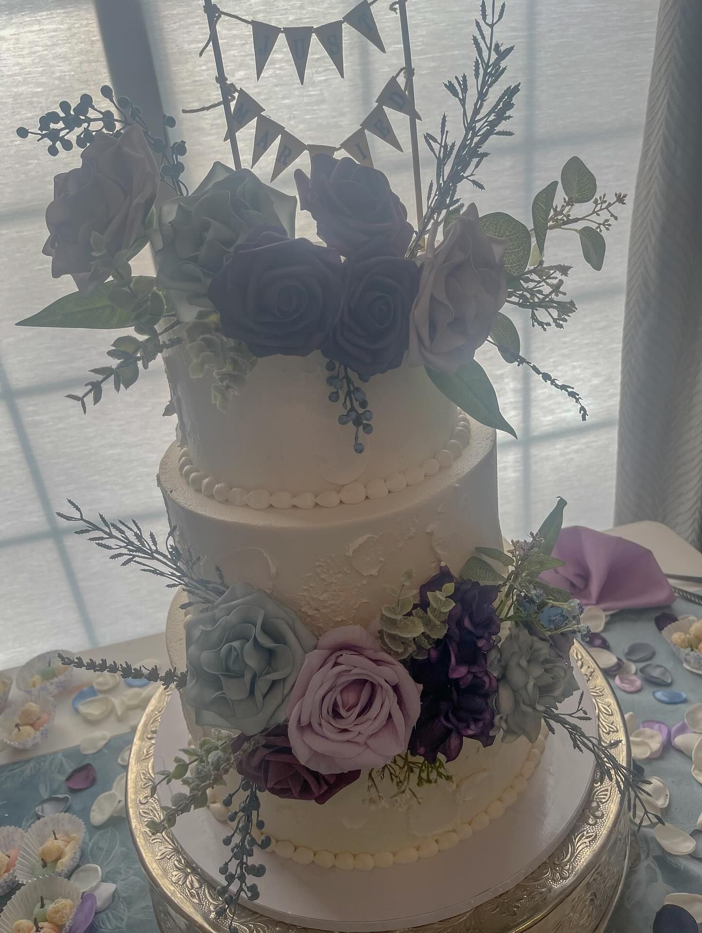 Three-tiered wedding cake, decorated with flowers in purple and green hues, topped with a banner.