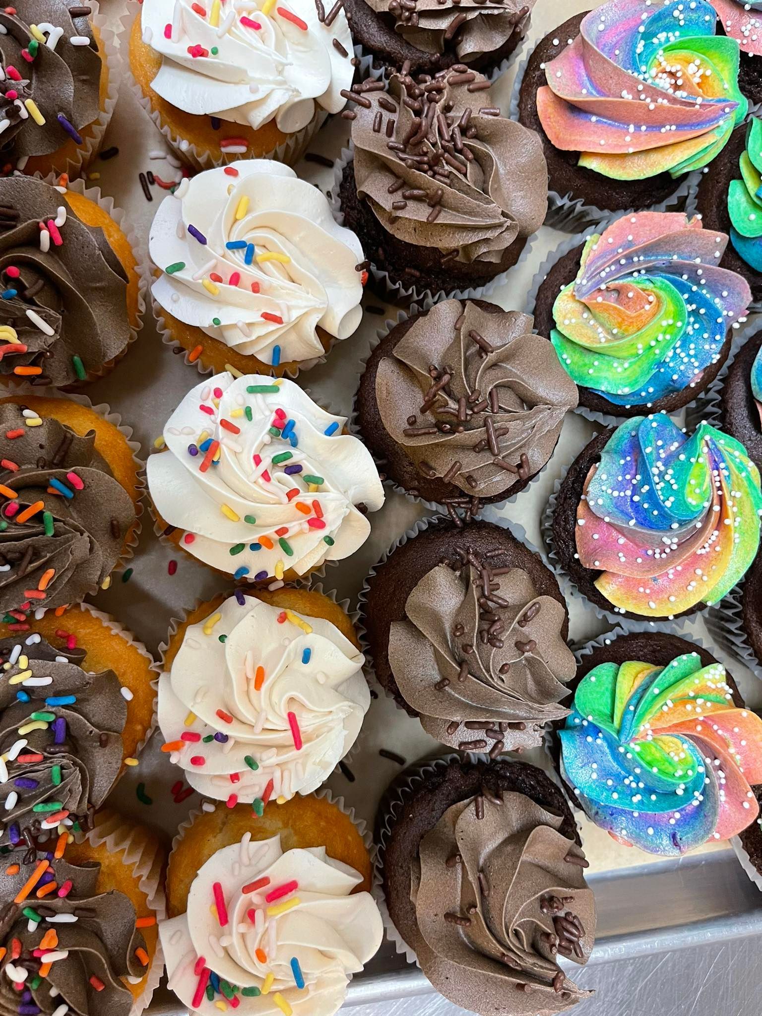 Cupcakes with various frostings and sprinkles on a tray; some are rainbow-swirled.