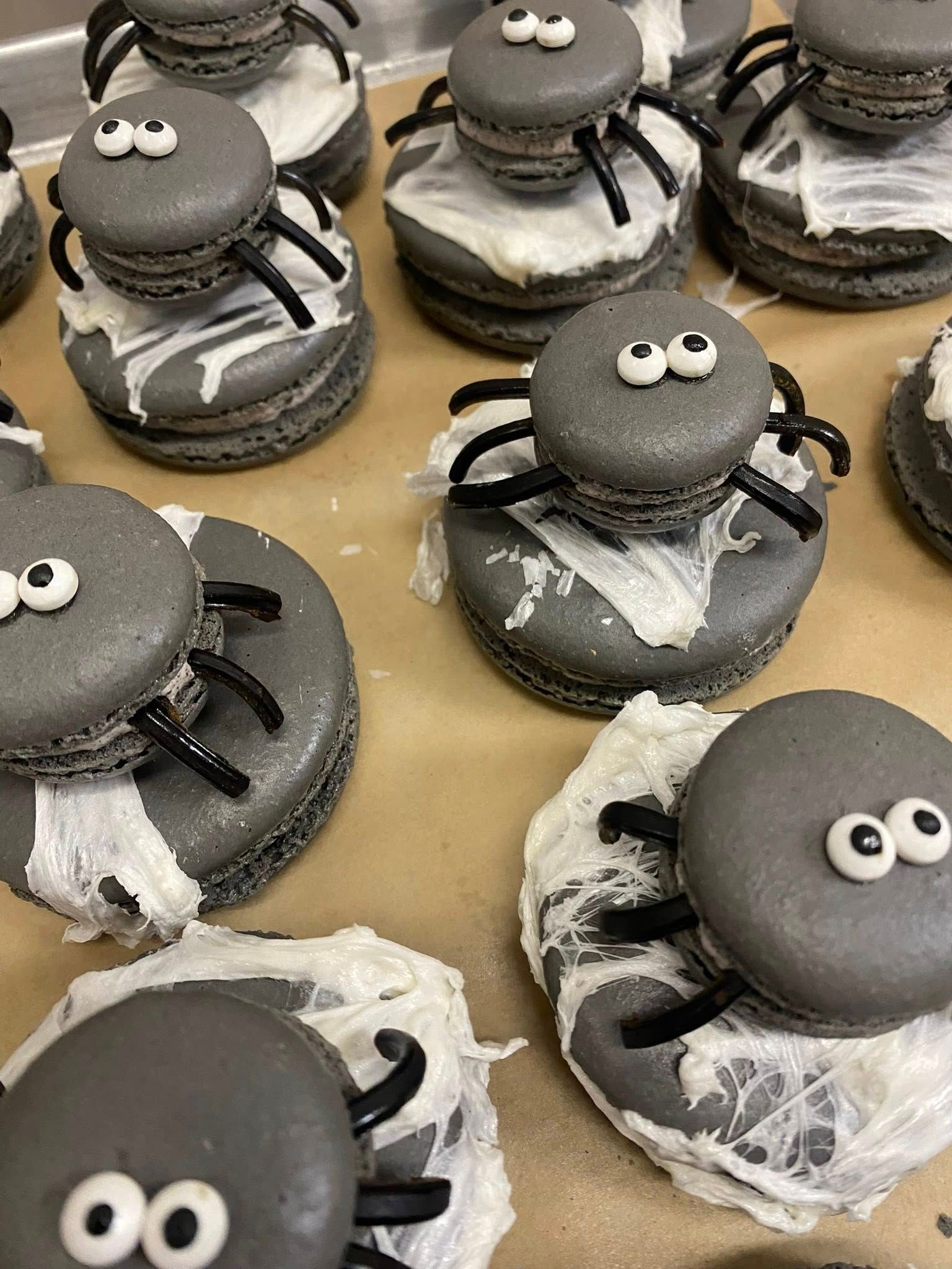 Gray macaron spider treats with candy eyes and licorice legs on a baking sheet, with sugar web decorations.