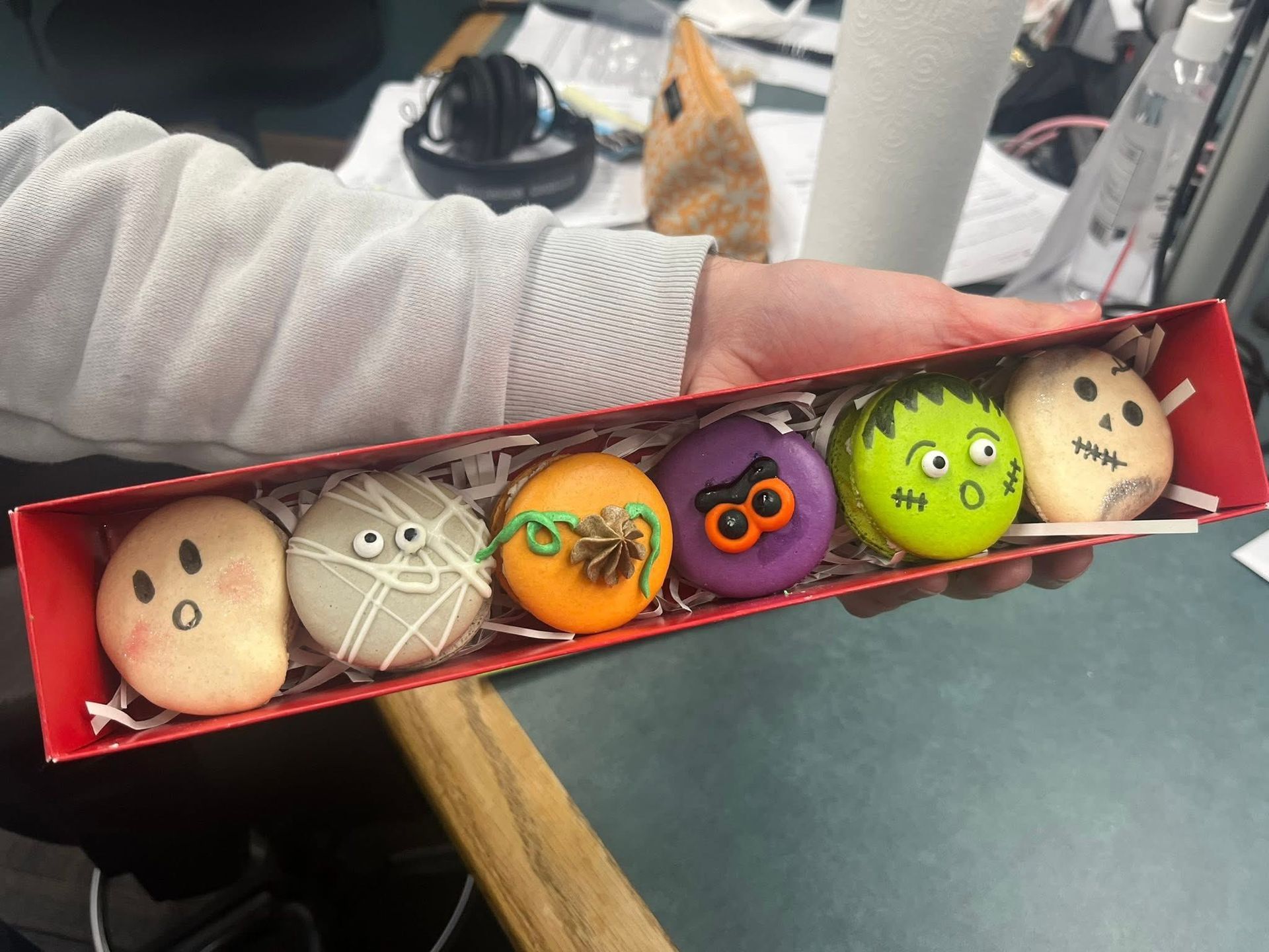 Person holding a red box of Halloween-themed macarons: ghost, spider, pumpkin, bat, Frankenstein, skull.