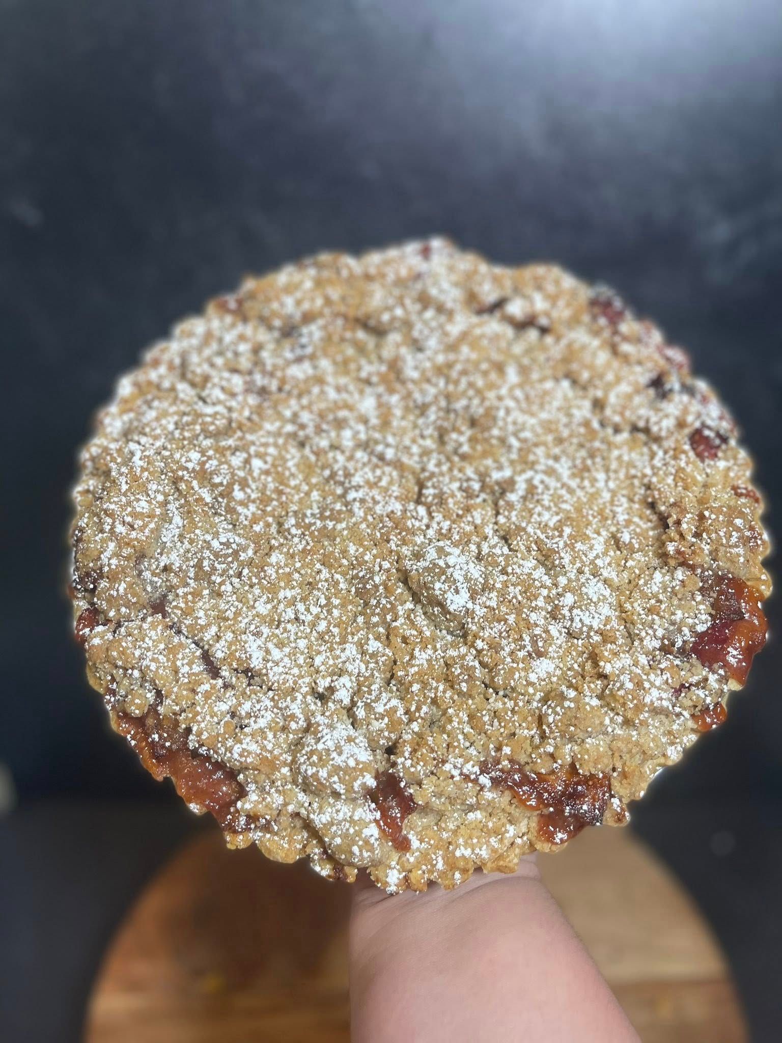Person holding a round, baked dessert with a crumb topping, dusted with powdered sugar.