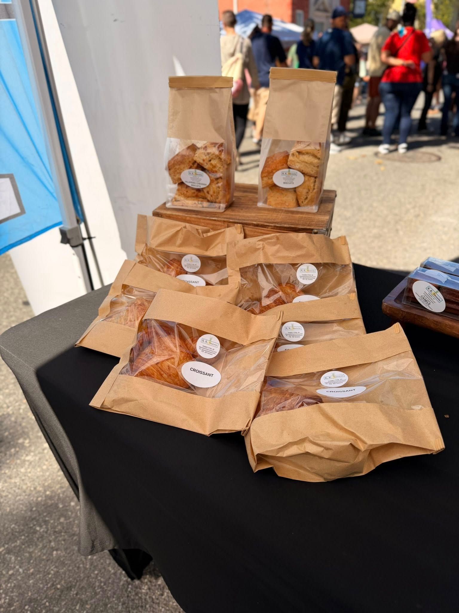 Brown paper bags of baked goods at an outdoor market on a black table; people in background.
