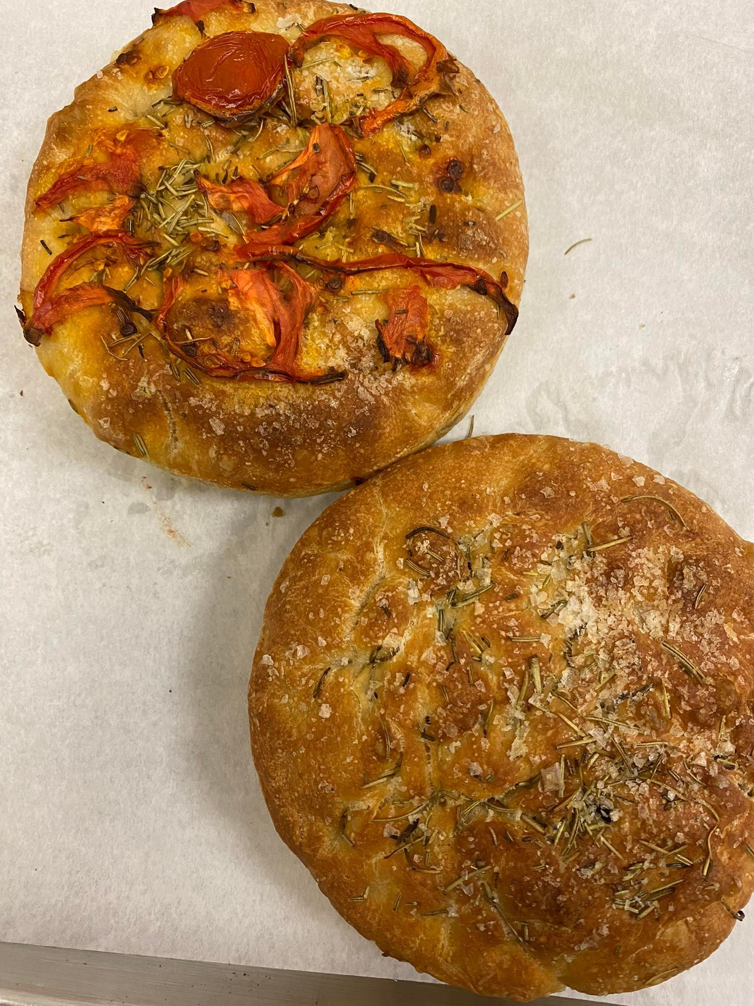 Two round focaccia breads, one with tomatoes and herbs, the other with herbs and cheese.