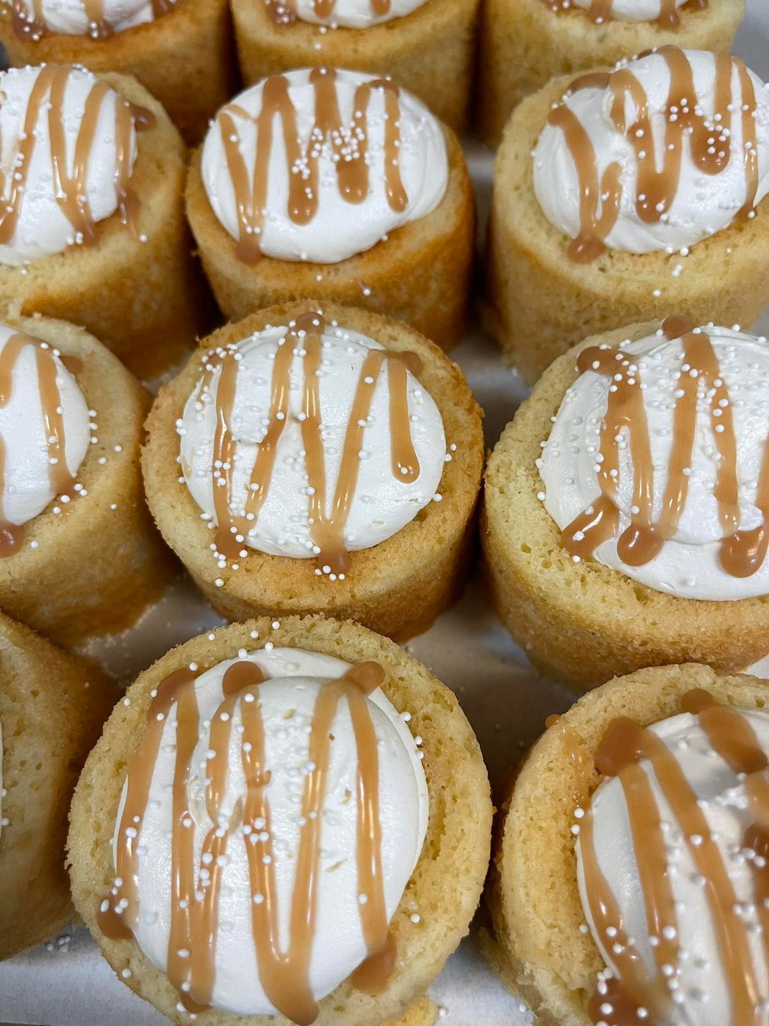 Close-up of caramel-topped cupcakes with white frosting and sprinkles in a light-colored setting.