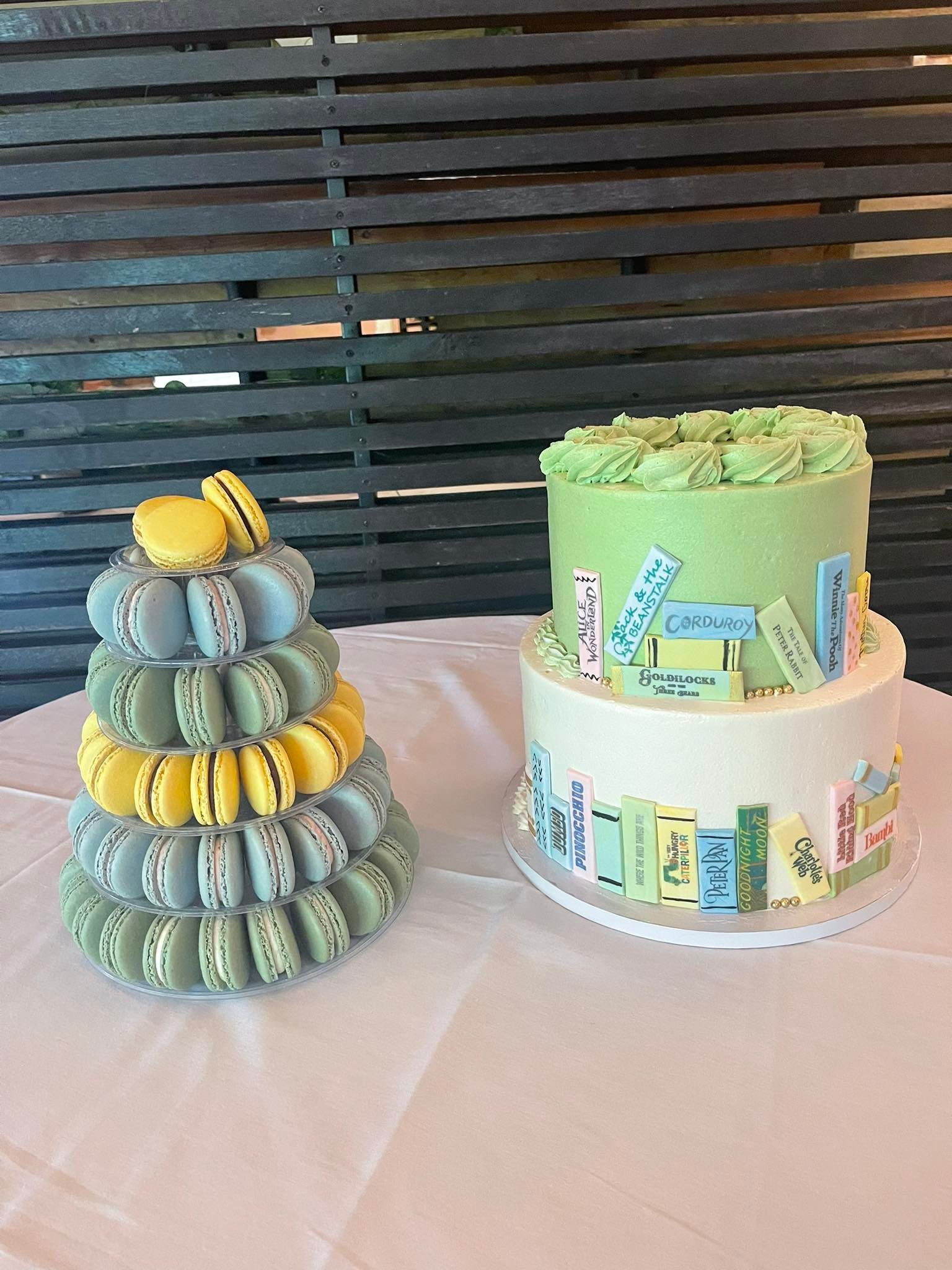 Macaron tower and book-themed cake on a white tablecloth, against a slatted wooden backdrop.