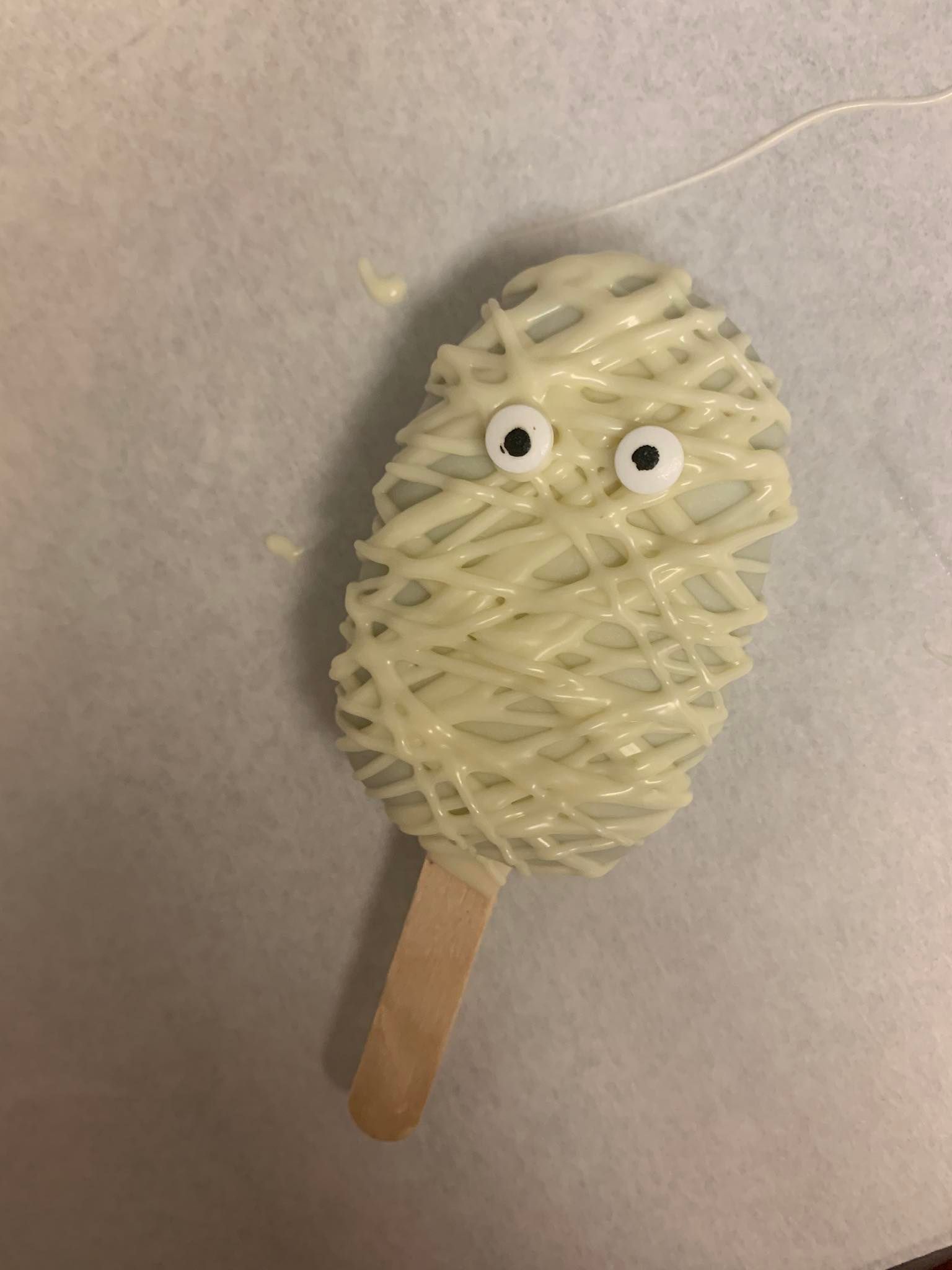 White chocolate-covered mummy popsicle with candy eyes on a light-colored surface.