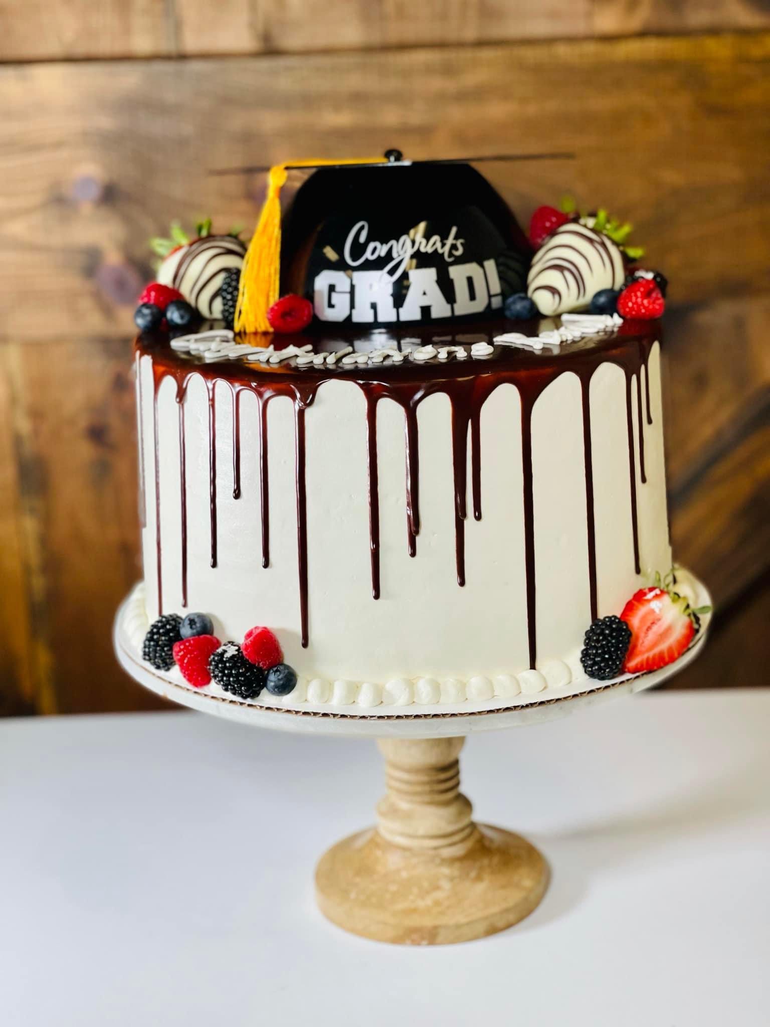 Graduation cake with white frosting, chocolate drip, berries, and a graduation cap on a wooden cake stand.