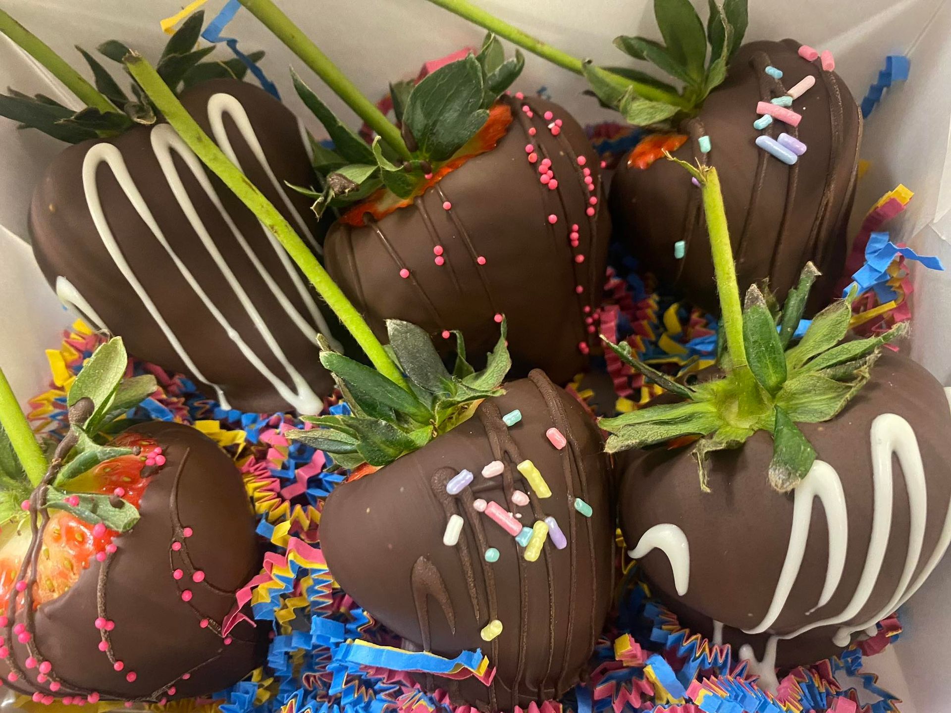 Chocolate-covered strawberries with white drizzle and sprinkles, arranged in a box with colorful paper.