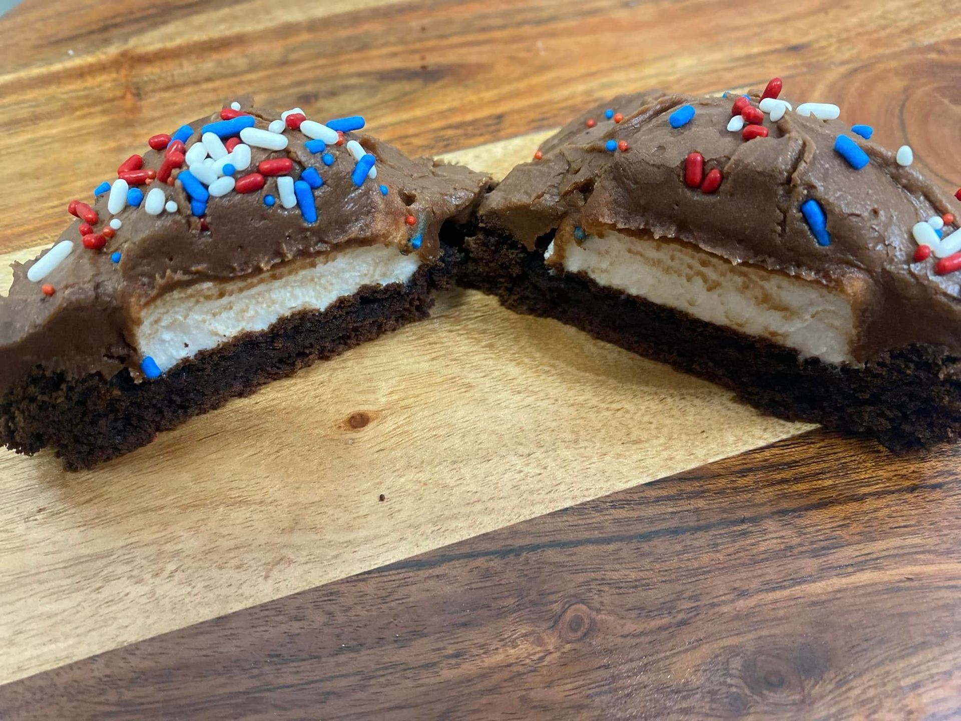 Two brownie halves with chocolate frosting, patriotic sprinkles, and a white center, on a wood surface.