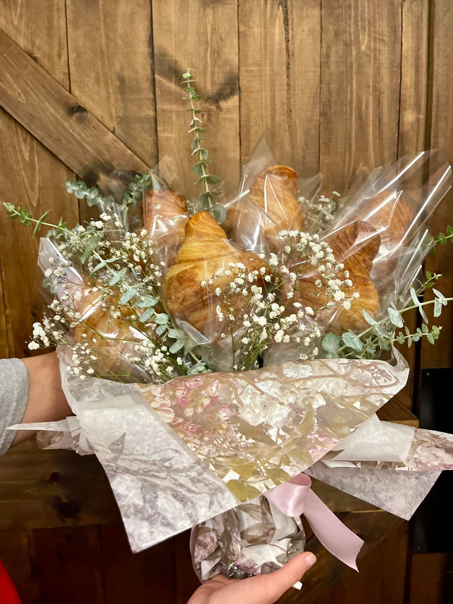A bouquet of wrapped croissants with baby's breath and a pink ribbon, held in front of a wooden door.