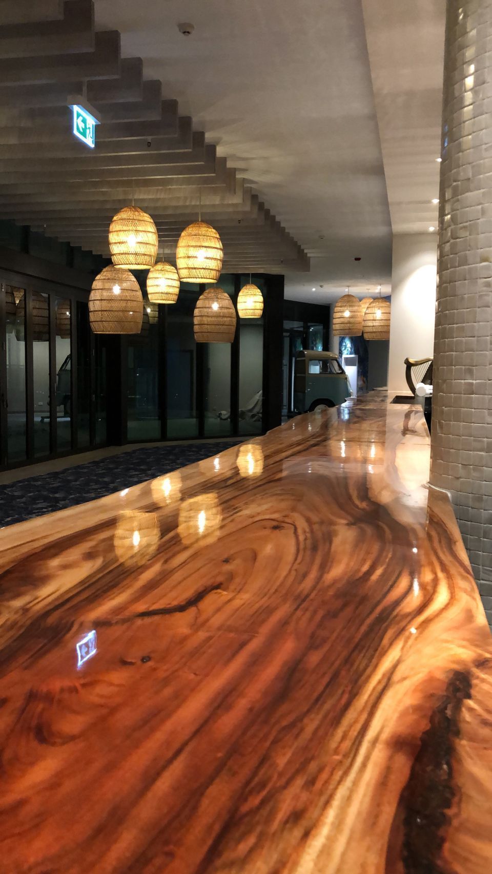 A glossy, polished wooden bar counter reflects warm pendant lights hanging from a modern, industrial-style ceiling.