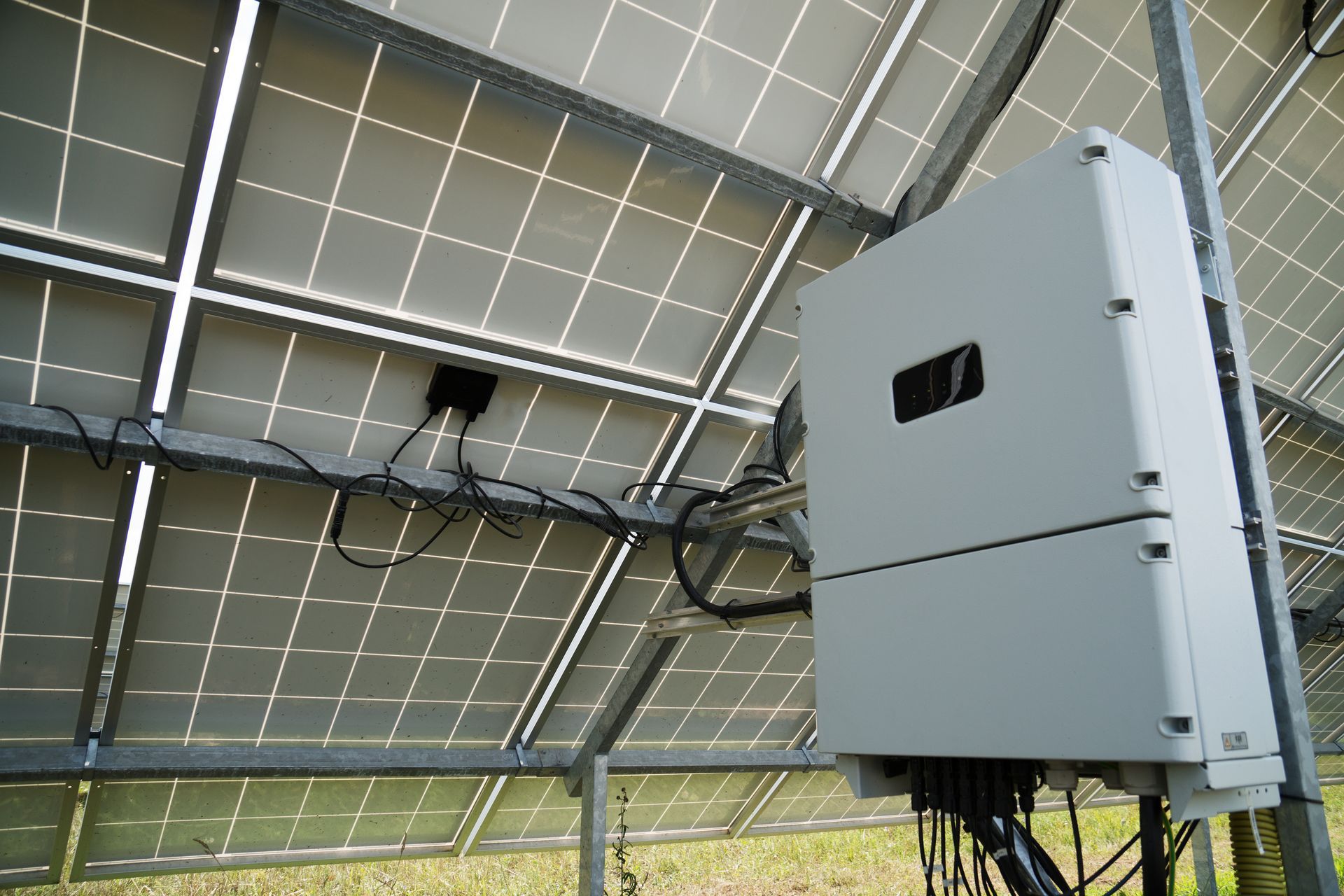 A gray electrical inverter mounted on a metal frame behind solar panels in a grassy field.