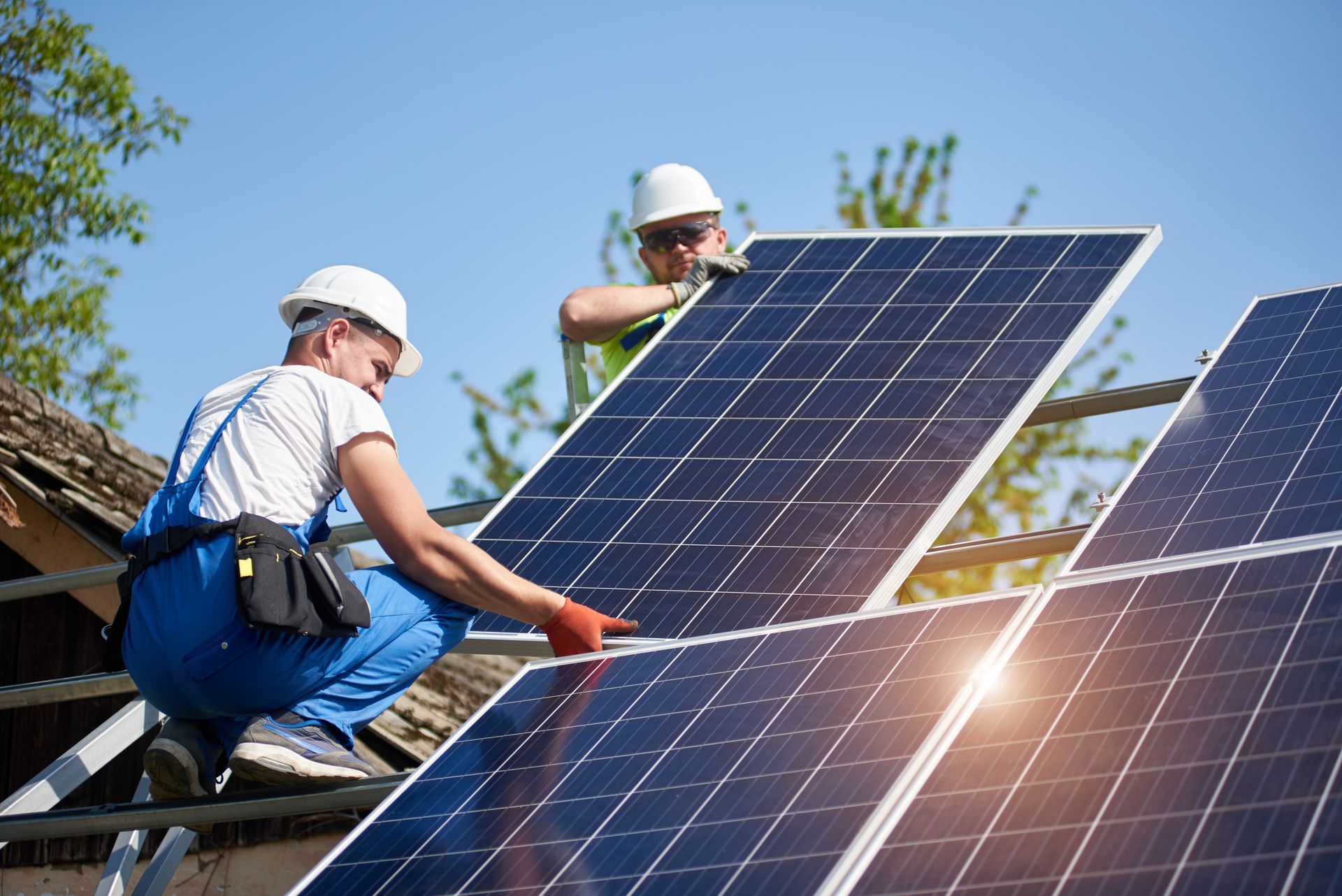 Two workers in hard hats and workwear install solar panels on a sunny, outdoor roof.