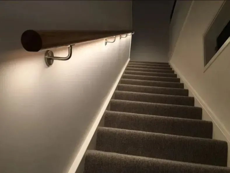 A wooden stair handrail mounted to a wall, illuminated from underneath by a soft, warm LED strip light.