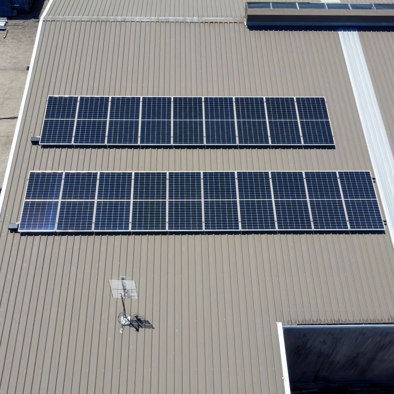 Two rows of solar panels installed on a beige corrugated metal roof, with an antenna and a roof hatch nearby.