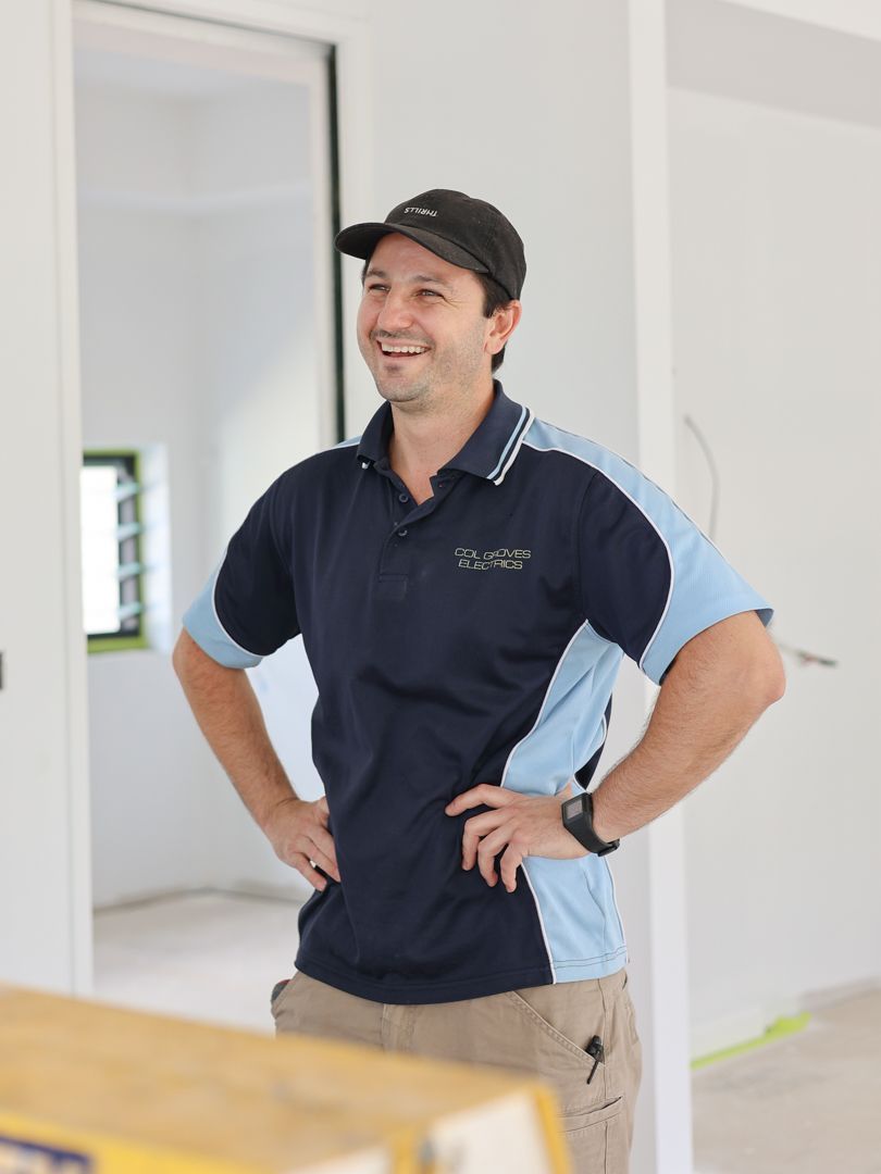A person wearing a black cap, a dark blue and light blue polo shirt, and khaki pants stands with hands on hips, smiling.