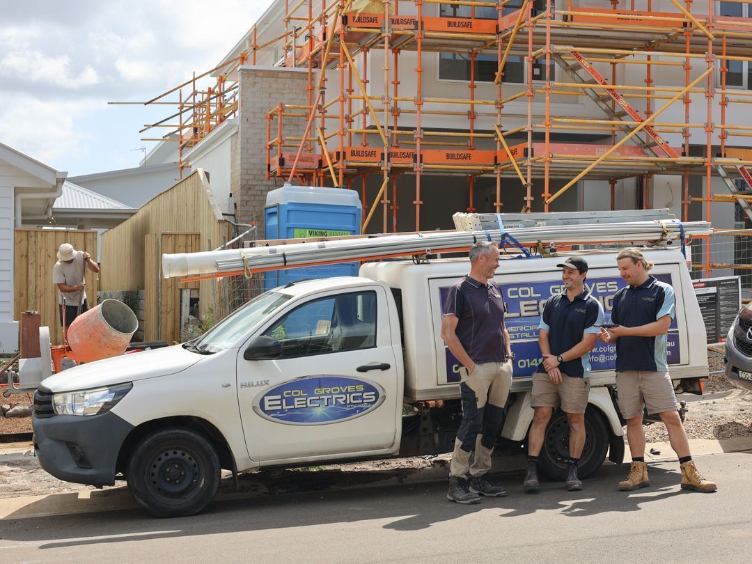 Three workers stand by a white trade vehicle in front of a construction site with scaffolding.