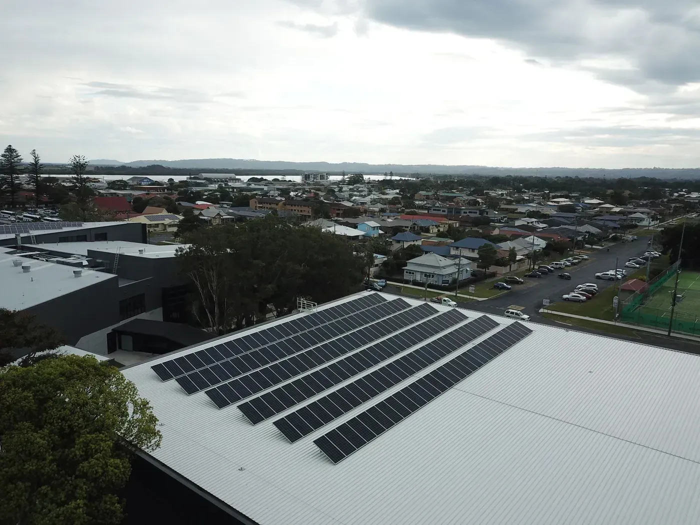 An aerial view shows a commercial building rooftop with large solar panel arrays overlooking a suburban town.