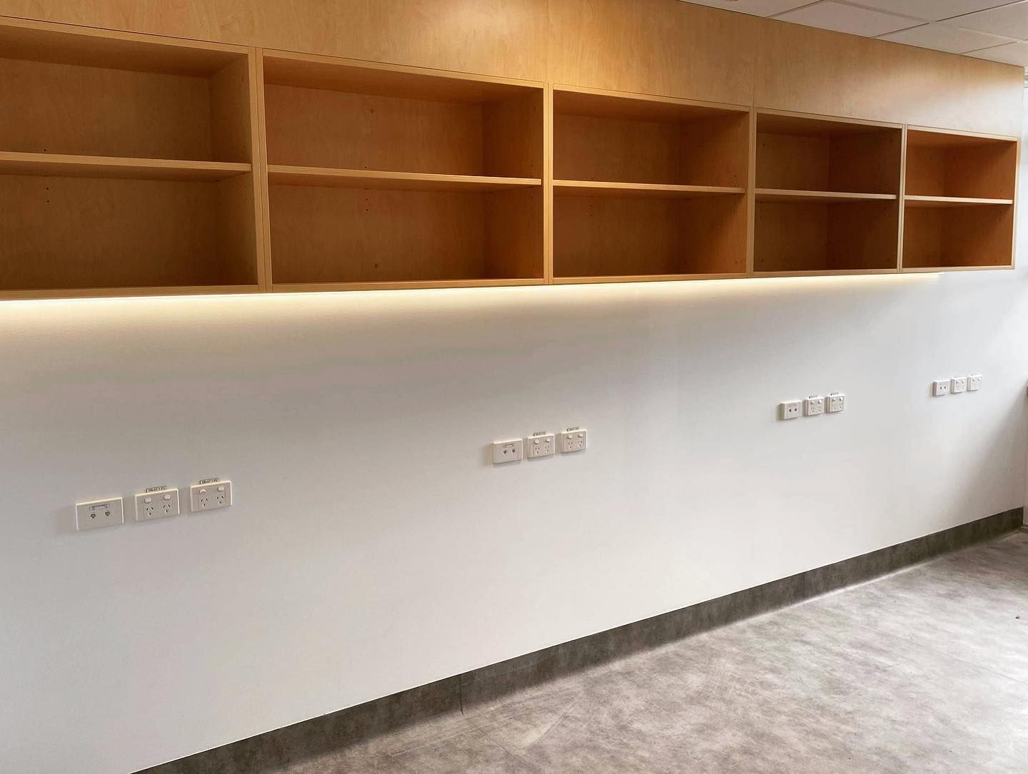 Empty light wood shelving mounted on a white wall above a series of electrical outlets.