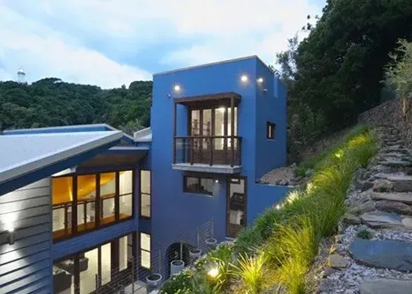 A modern two-story blue house with a balcony, set against a hillside with a stone staircase and outdoor garden lighting.