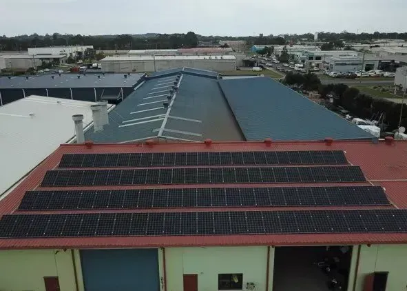 Aerial view of a red-roofed warehouse building featuring several rows of dark solar panels installed on its roof.