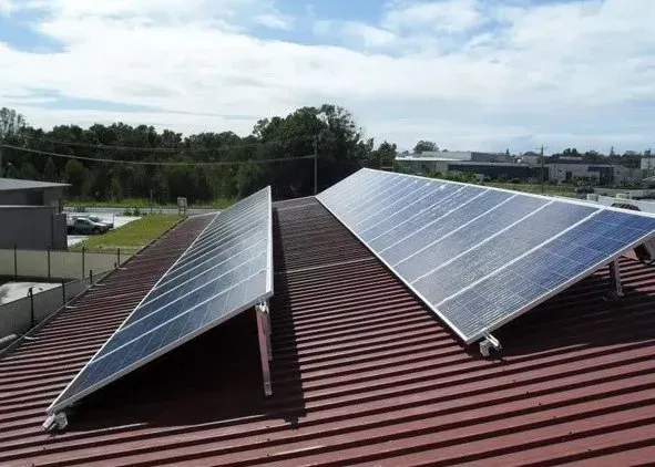 Two rows of solar panels installed on a red corrugated metal roof under a bright, cloudy sky.