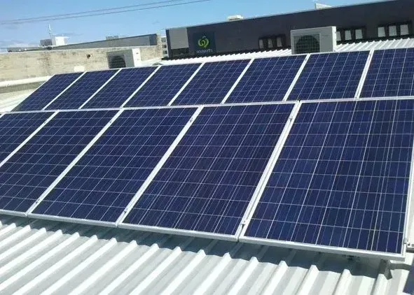 A grid of dark blue rectangular solar panels installed on a white corrugated metal roof under a bright blue sky.