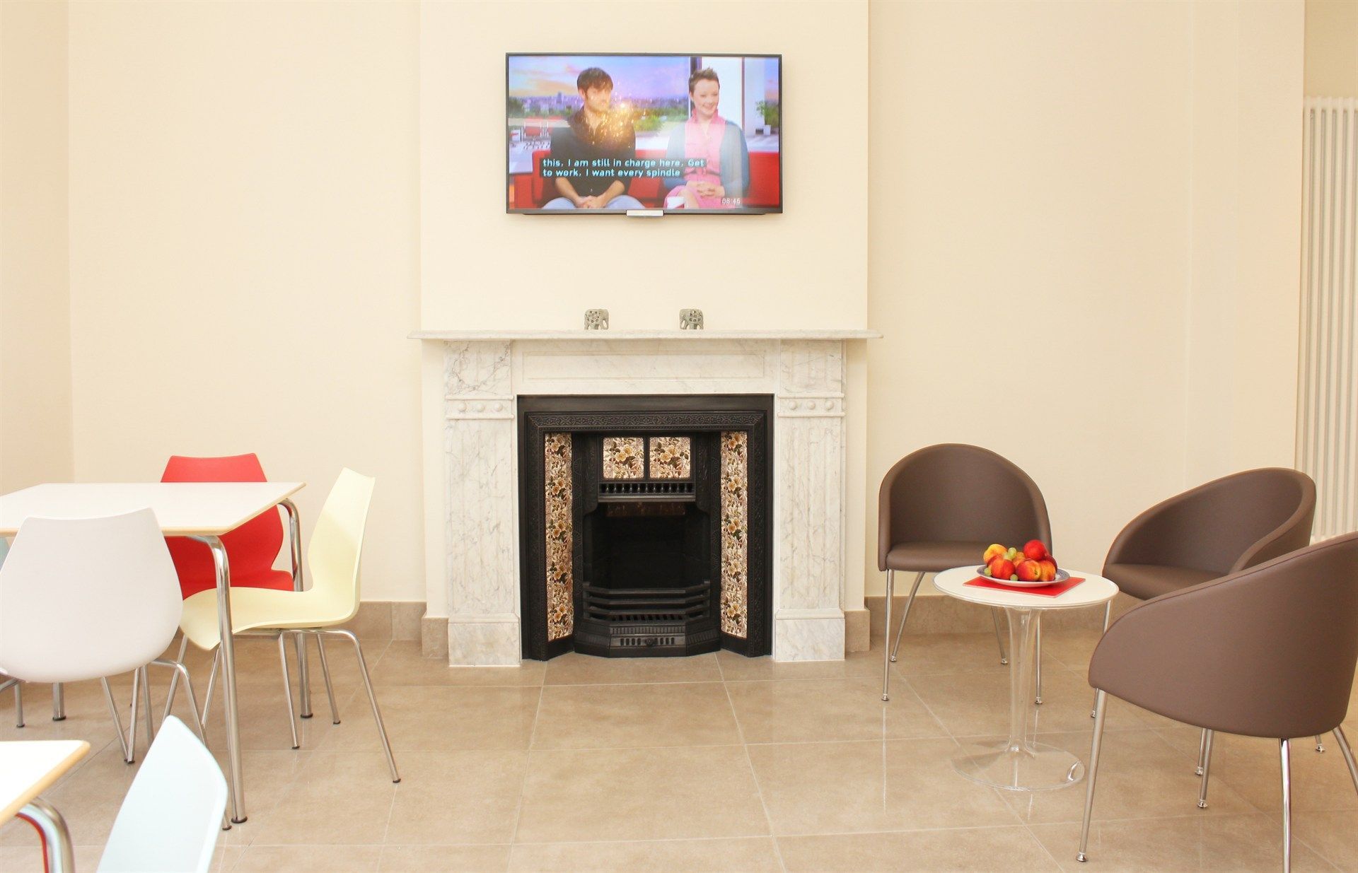 A living room with a fireplace , chairs , tables and a flat screen tv.
