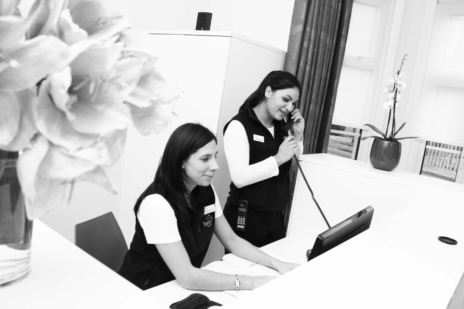 Two women are sitting at a desk talking on a phone