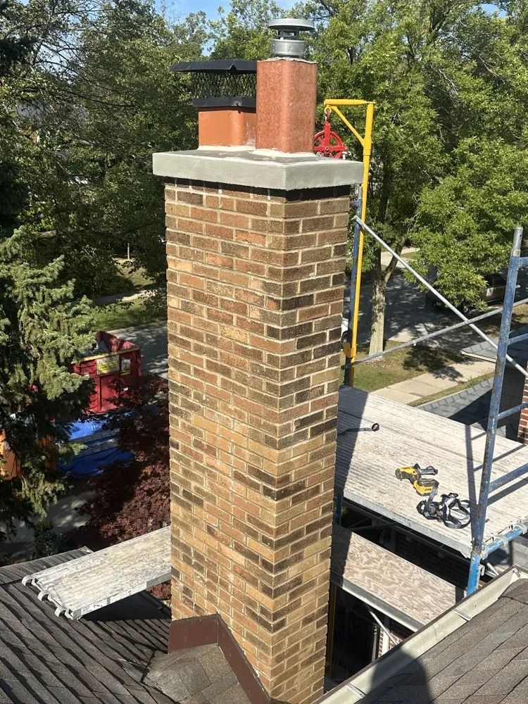 Brick chimney on a roof with a gray cap, surrounded by trees and scaffolding.