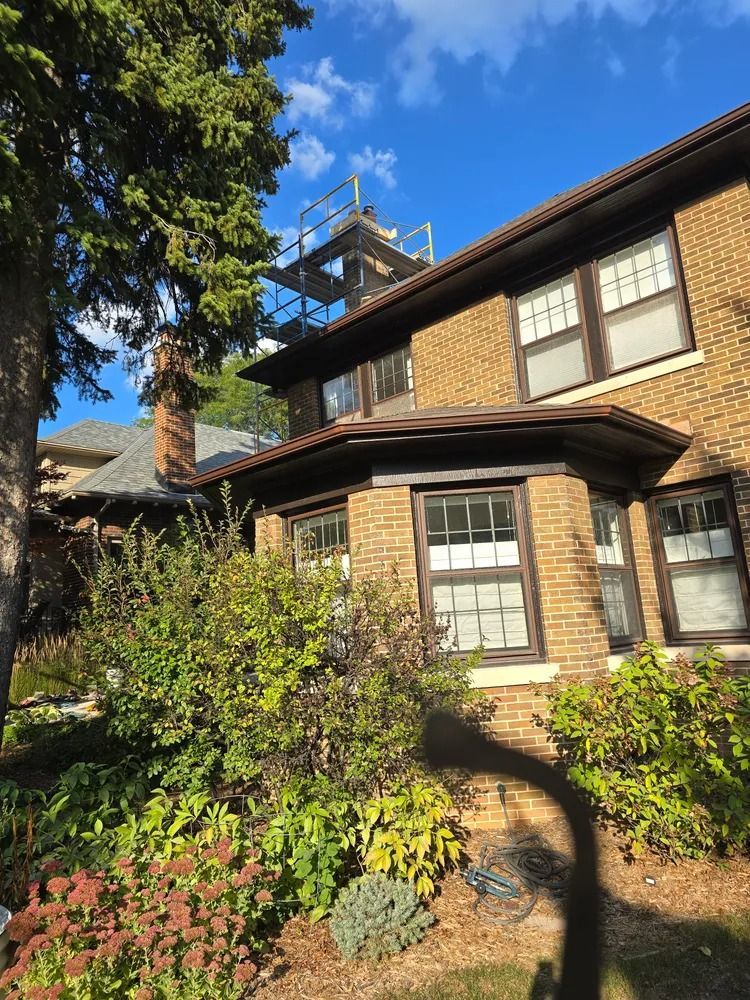 Brick house with dark trim and scaffolding on the roof. Bushes and trees in front on a sunny day.