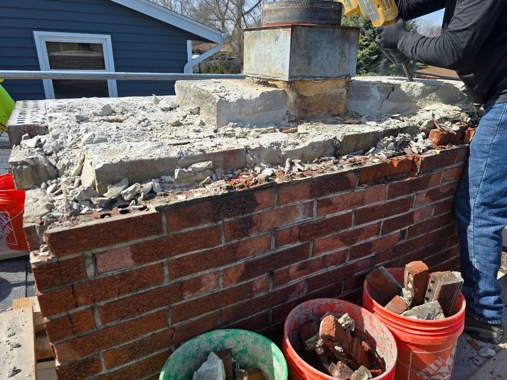 Person demolishing a brick chimney with a jackhammer; broken bricks and rubble in buckets.