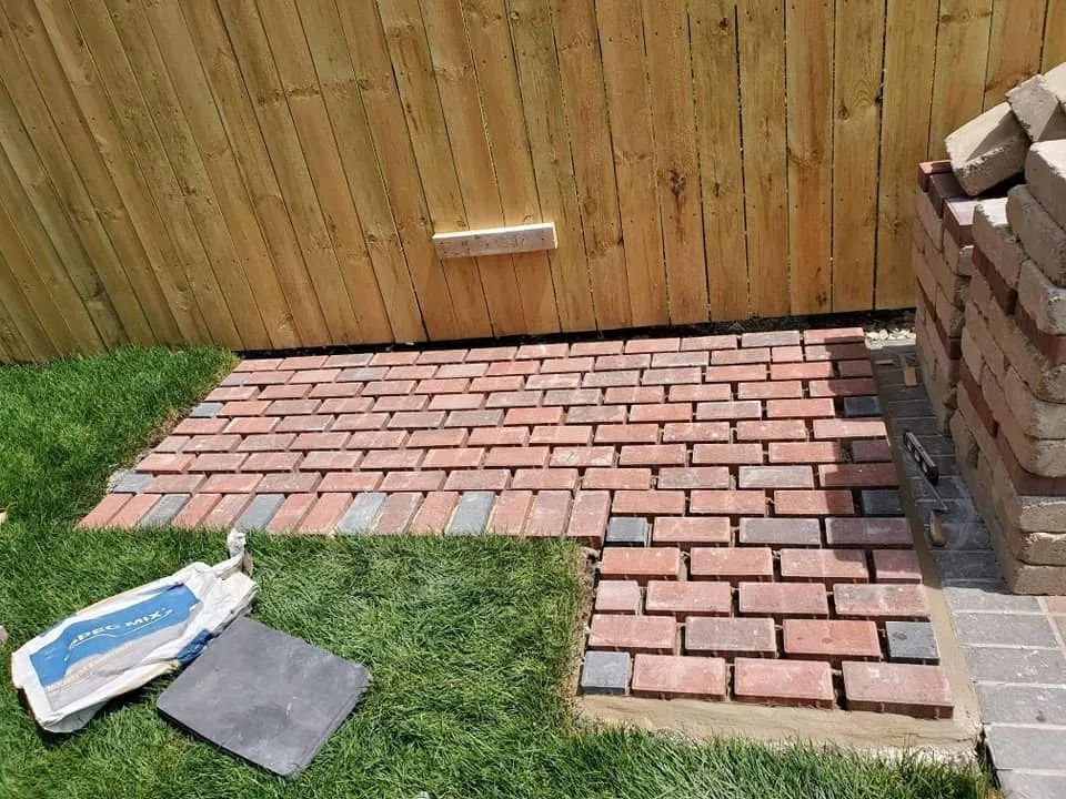 Brick patio being constructed next to a wooden fence on grass; bricks in red, gray, and brown.