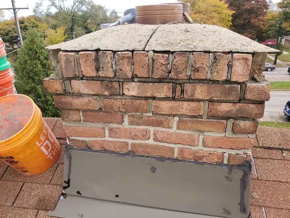 Chimney with brickwork, concrete cap, and flashing on a roof with orange buckets nearby.